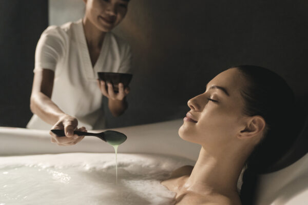 Woman relaxing in a therapeutic balneotherapy while a spa staff member adds seaweed extract, demonstrating one of treatments available at Chiva-Som Hua Hin